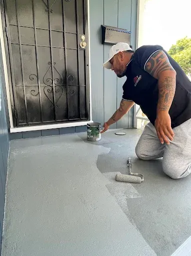 Man painting a porch gray, near a black door and mailbox. He kneels, holding a paint can and roller.