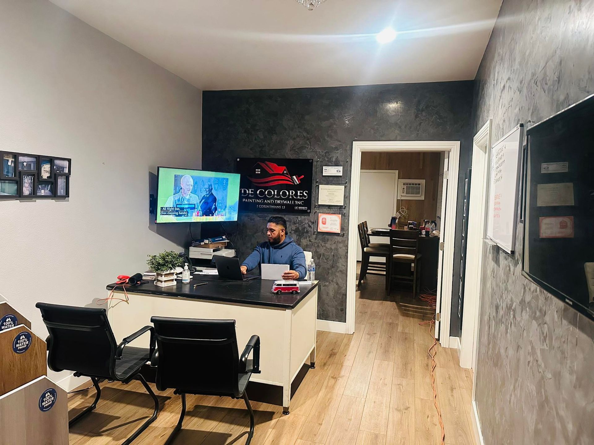 Man at desk in office, two chairs, TV, gray walls, door leads to other room.