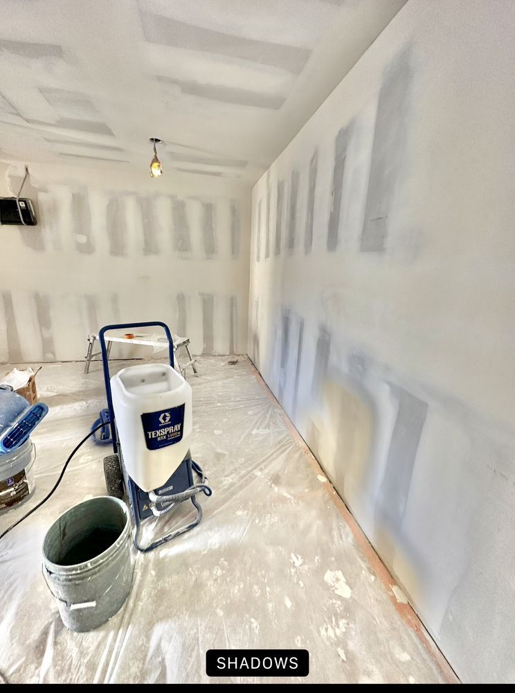 Interior wall with drywall being worked on, showing paint sprayer and other equipment on a covered floor.