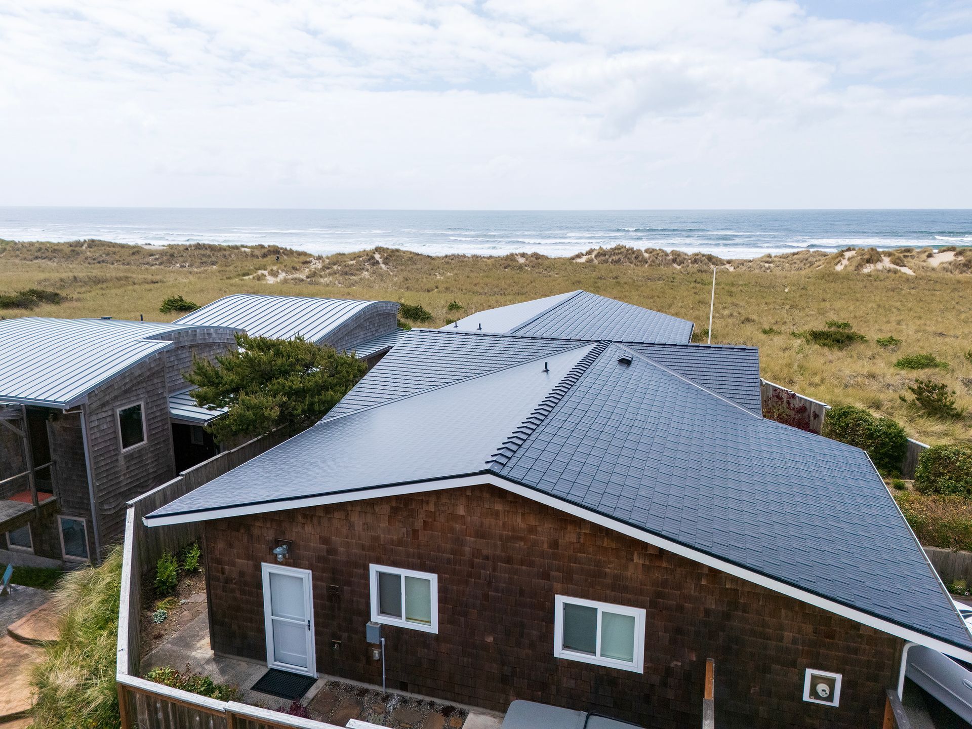 Side perspective of a Manzanita Beach residence showcasing a new black aluminum Interlock Cedar Shingle Roof, installed by Interlock Metal Roofing in the Buena Vista neighborhood of Tillamook County, Oregon. The home’s shingle-style façade is complemented by energy-efficient windows and a private courtyard walkway. Designed to endure frequent wind gusts exceeding 70 mph, the 4-way interlocking metal shingles create a seamless, watertight surface ideal for coastal living. The corrosion-resistant aluminum construction and Alunar® protective coating defend against the harsh marine environment and wind-driven rain.