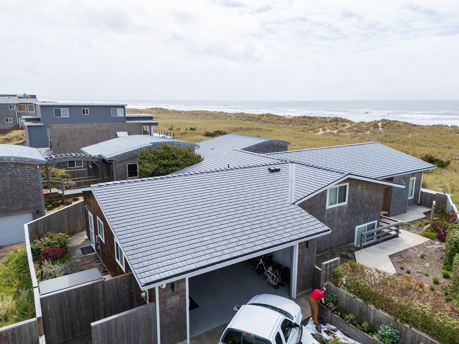 Overhead view of coastal property located steps from Manzanita Beach in the Buena Vista neighborhood, featuring a high-performance Black Interlock Cedar Shingle Roof. Installed by Interlock Metal Roofing, the durable aluminum roofing system offers a sleek aesthetic and is optimized for extreme coastal wind conditions. The 4-way interlocking panel system ensures roof panels remain securely fastened during windstorms and resists salt-laden air thanks to Alunar® coating technology. The architecture highlights include a low-pitched gable roof, weathered cedar siding, and double-pane vinyl windows, all oriented toward the oceanfront dunes.