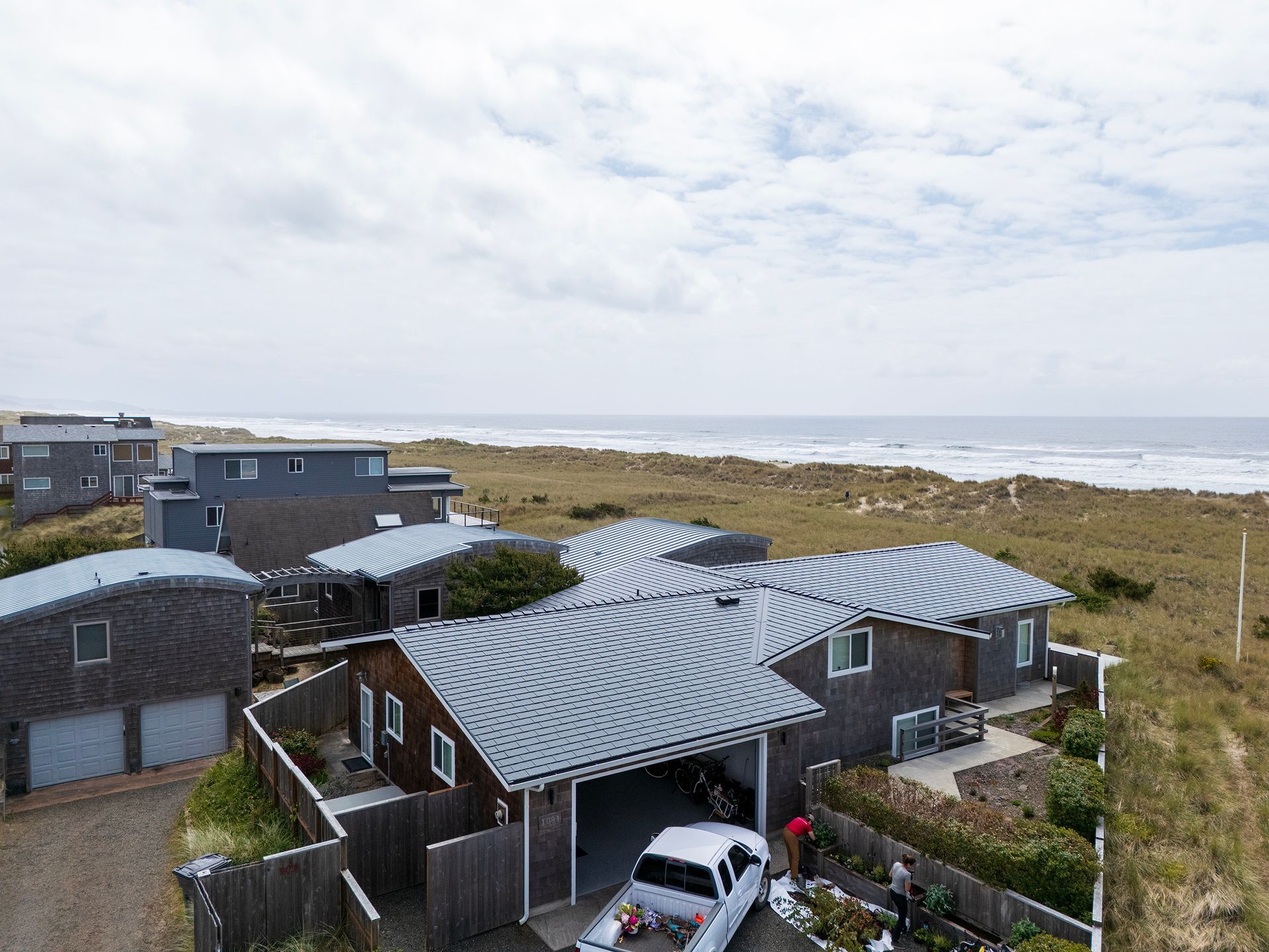 Wide-angle aerial view of an oceanfront home in Manzanita’s Buena Vista neighborhood, clad in natural cedar shake siding and topped with a Black Interlock Cedar Shingle aluminum roof. Installed by Interlock Metal Roofing, the heavy-duty system is fortified with a 4-way interlocking mechanism ideal for Oregon Coast wind conditions. Featuring integrated ridge caps, salt spray resistance, and a fade-resistant Alunar® finish, the roofing blends form and function. The landscape includes fenced perimeter gardens and a dual-gabled entryway with covered porch and attached garage.