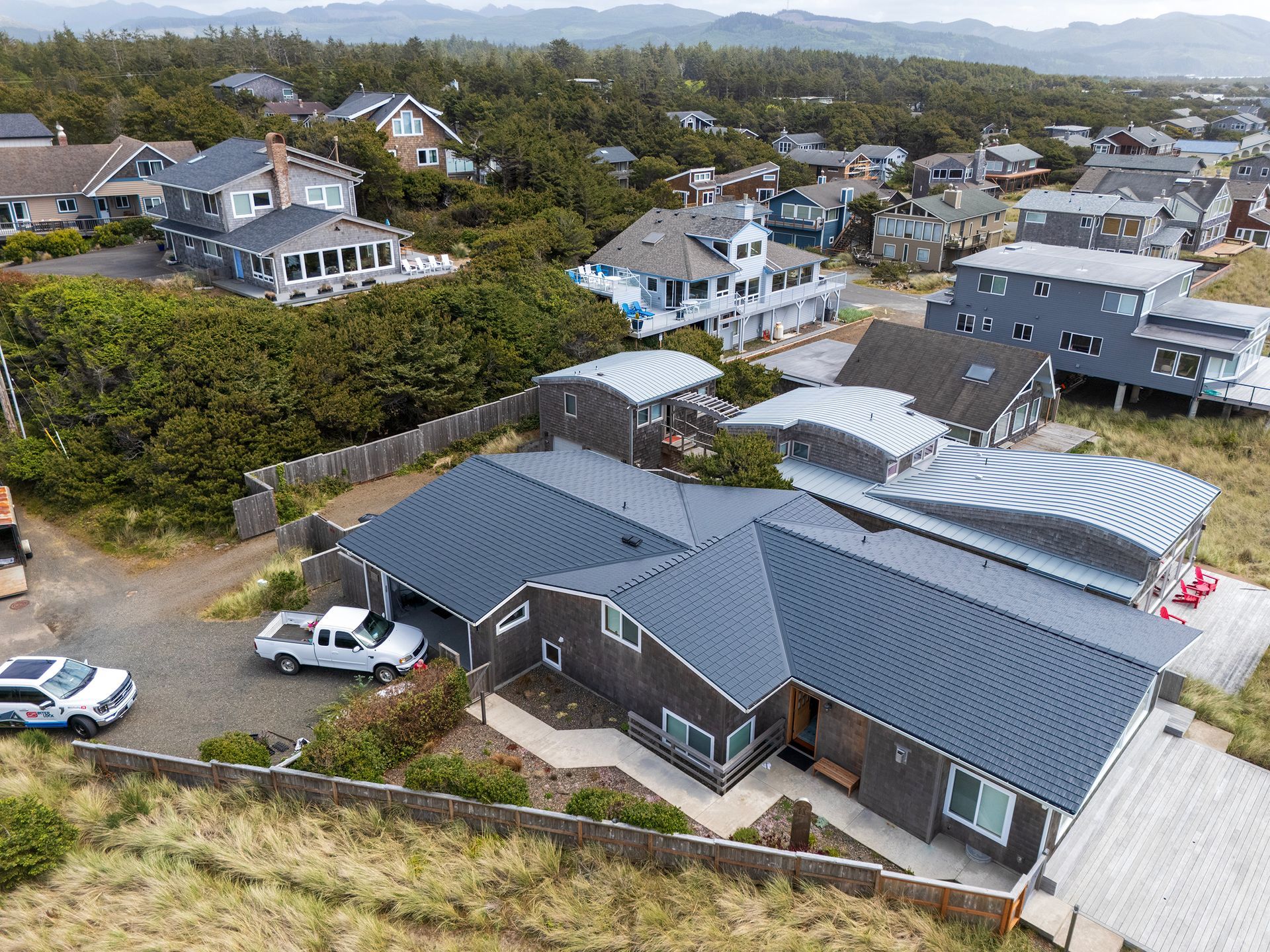 Front aerial view of a custom coastal home in the Buena Vista neighborhood of Manzanita, Oregon, showcasing a Black Interlock Cedar Shingle Roof made from heavy-gauge aluminum with a 4-way interlocking system for extreme wind protection. The low-pitch gable roofline spans multiple wings of the house, highlighting the matte-finished shingles that resist corrosion from oceanfront salt spray, thanks to the durable Alunar® Coating. Surrounded by sand dunes and coastal grasses near Manzanita Beach, this home features rustic cedar shake siding, white-trimmed energy-efficient windows, and modern entryway accents beneath a highly wind-resistant aluminum roof installed by Interlock Metal Roofing.