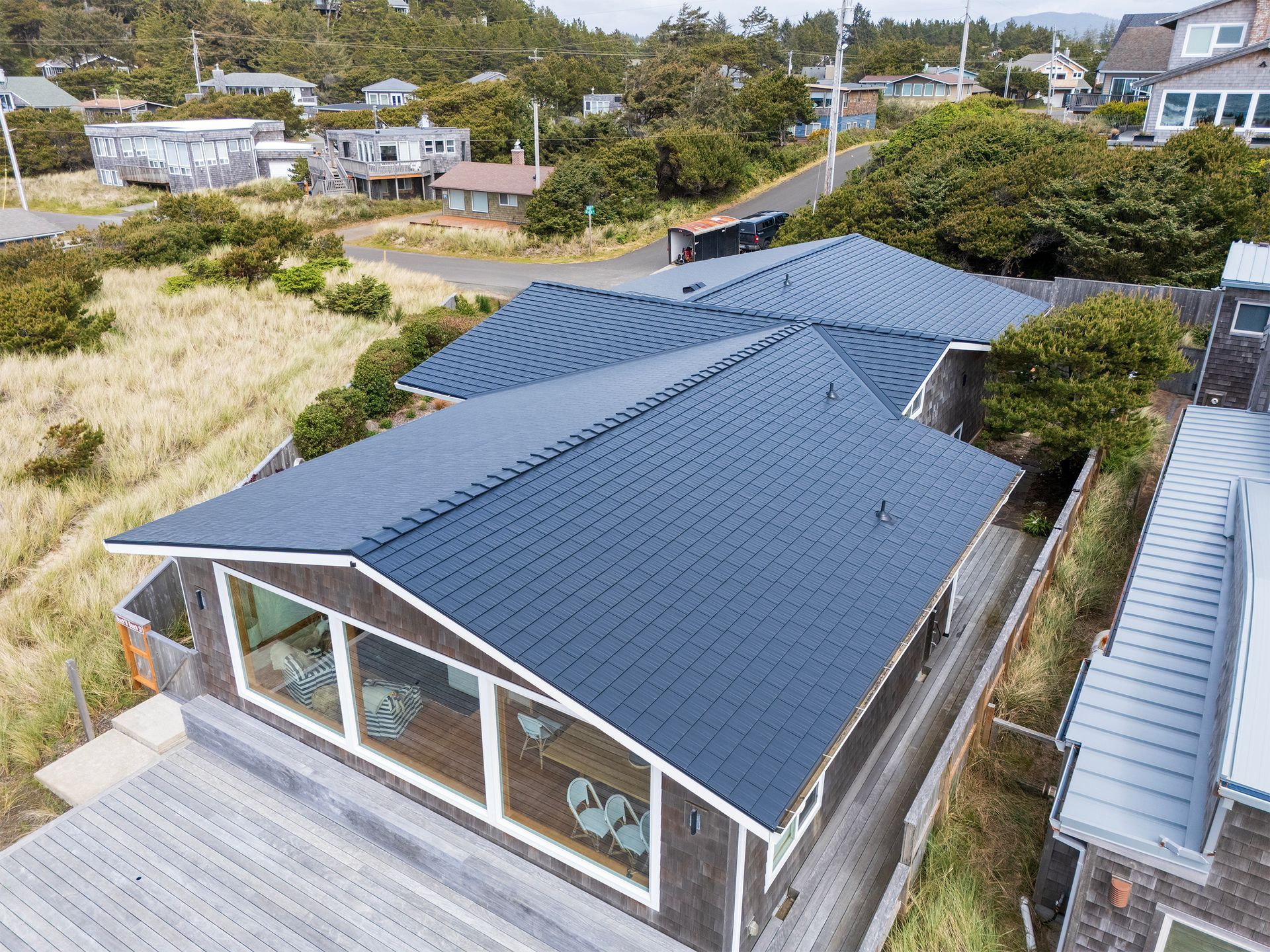 Oblique rear angle of a Manzanita Beach home fitted with a premium Black Interlock Cedar Shingle Roof, engineered with heavy-gauge aluminum and a 4-way locking system for hurricane-strength wind uplift resistance. Set in the oceanfront Buena Vista neighborhood, this Oregon coast property features full-length clerestory windows beneath a sweeping gabled roofline that maximizes views and sunlight. The sleek black shingles offer the aesthetic of natural cedar but provide lifetime durability, rust resistance, and salt spray protection — essential for homes just steps from the Pacific Ocean. Installed by Interlock Metal Roofing for high performance in coastal environments.