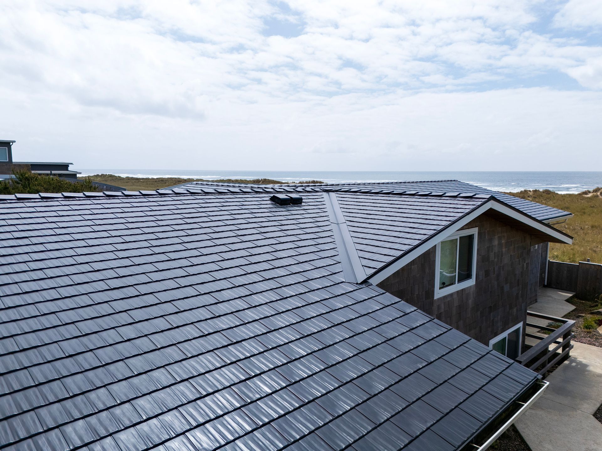 Close-up of a gleaming Black Interlock Cedar Shingle aluminum roof installed on a coastal Oregon home by Interlock Metal Roofing. Located directly on Manzanita Beach in Tillamook County’s Buena Vista neighborhood, this roof highlights the strength of its 4-way interlocking design, which prevents shingle uplift and leakage in high-wind events. The premium Alunar® coating technology shields against corrosion and sea salt intrusion, ensuring long-term protection and minimal maintenance. The architecture includes modern gable lines, white-trimmed windows, and a multi-level cedar plank walkway.