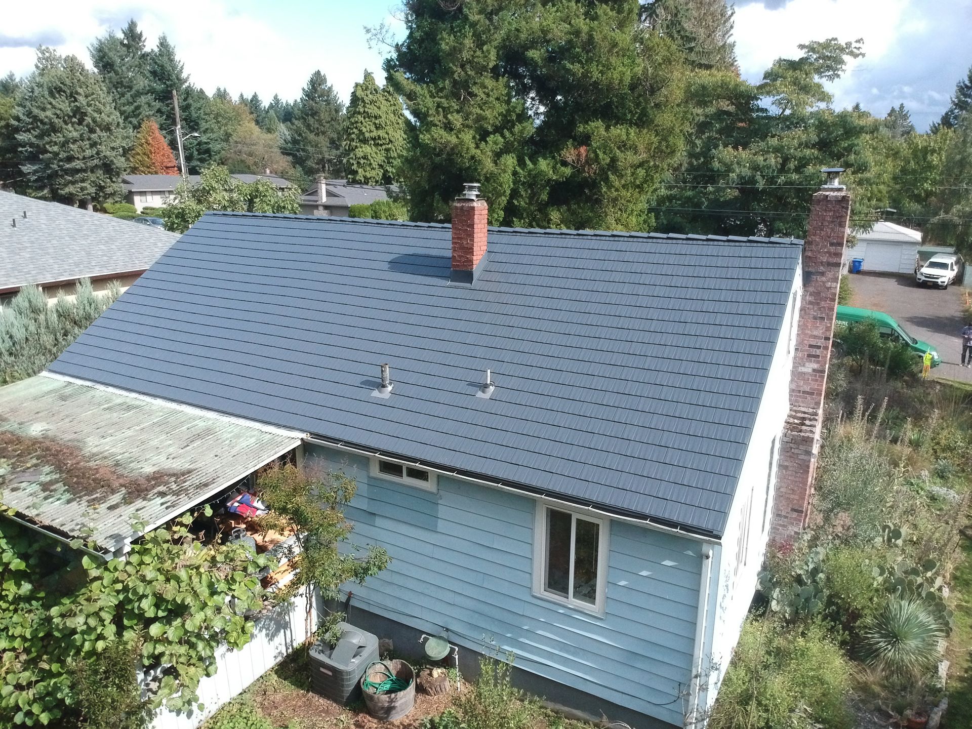 Aerial view of a charming single-story craftsman-style home in the Hazelwood neighborhood of Portland, Oregon, featuring a newly installed Deep Charcoal Interlock Cedar Shingle Roof made from heavy-gauge, durable aluminum by Interlock Metal Roofing. The roof showcases clean ridge lines and modern vent pipe flashings, contrasting beautifully with the home’s light blue horizontal wood siding. The house includes white-framed vinyl windows, a prominent red brick chimney, and a rustic shed with a weathered corrugated metal roof partially covered in overgrown vines. The property is landscaped with native Oregon greenery, including coniferous trees and desert plants like cactus and yucca, reflecting the Pacific Northwest climate. This unique Portland home exemplifies energy-efficient aluminum roofing and timeless architectural features, making it a standout installation by Interlock Metal Roofing in Multnomah County.