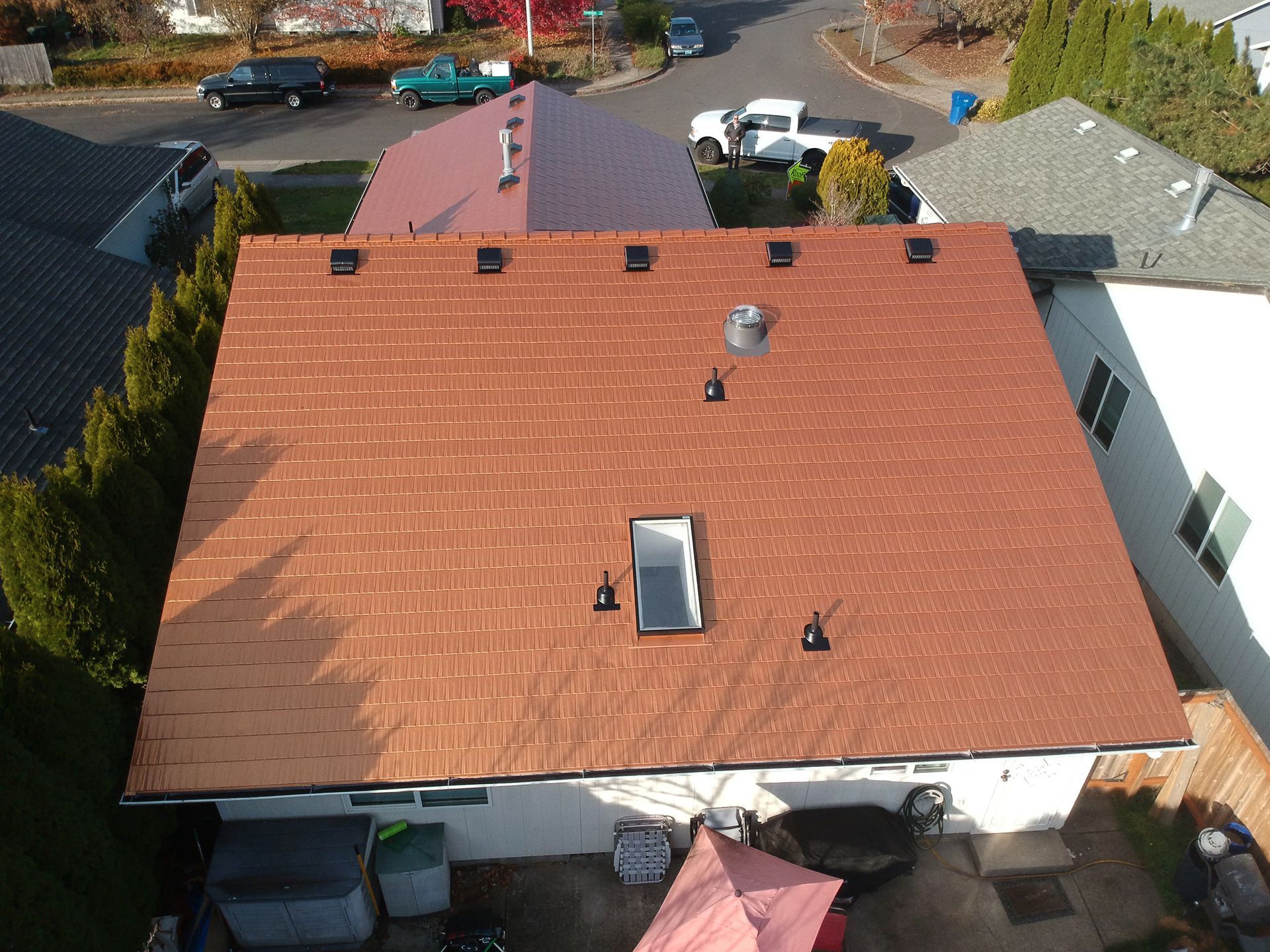 Top-down aerial view of a home in Southeast Mill Creek, Salem, Oregon, showcasing a freshly installed Copper Penny Interlock Cedar Shingle Roof by Interlock Metal Roofing. Designed to mimic the elegance of natural copper without oxidization or patina, the roof’s heavy gauge aluminum construction offers rust-free performance and long-term energy efficiency. Notable rooftop features include a low-profile solar tube skylight for passive daylighting, multiple factory-painted roof ventilation outlets for attic airflow, and a precision-fitted pipe vent flashing for leak protection. The copper-toned metal cedar shingles are coated with Interlock’s proprietary Alunar® coating for enhanced UV and weather resistance. Paired with the home’s rich wood siding, white rain gutters, and secluded forest-edge setting, this roof adds both curb appeal and resilience to the property.
