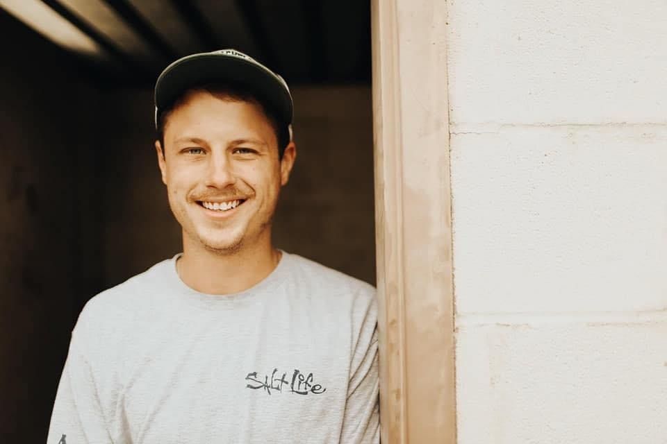 Man in a grey shirt and green cap smiles, leaning in a doorway with white block walls.