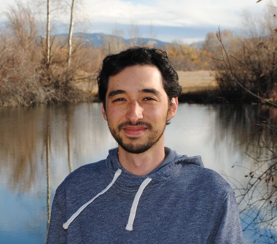 Man in blue hoodie smiles in front of a lake, trees, and a mountain range.