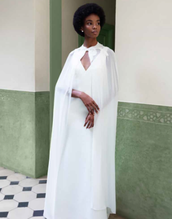 A woman in a white dress with a cape is standing in a hallway.