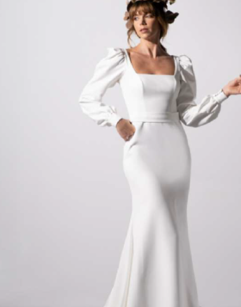 A woman in a white dress with puffed sleeves is standing in front of a white wall.