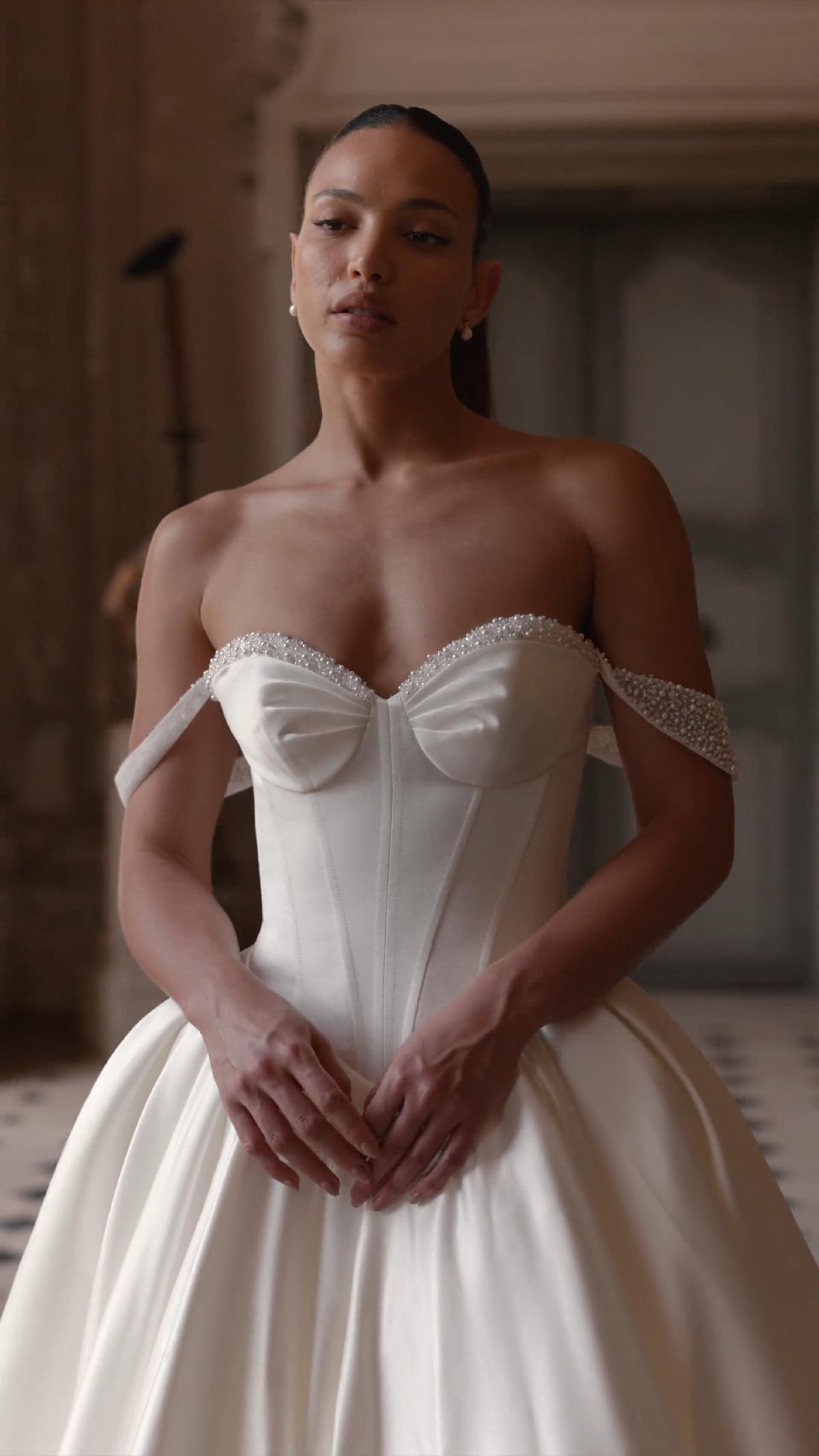 Woman in a white off-the-shoulder wedding gown. Standing indoors, arms crossed over chest, looking upward.
