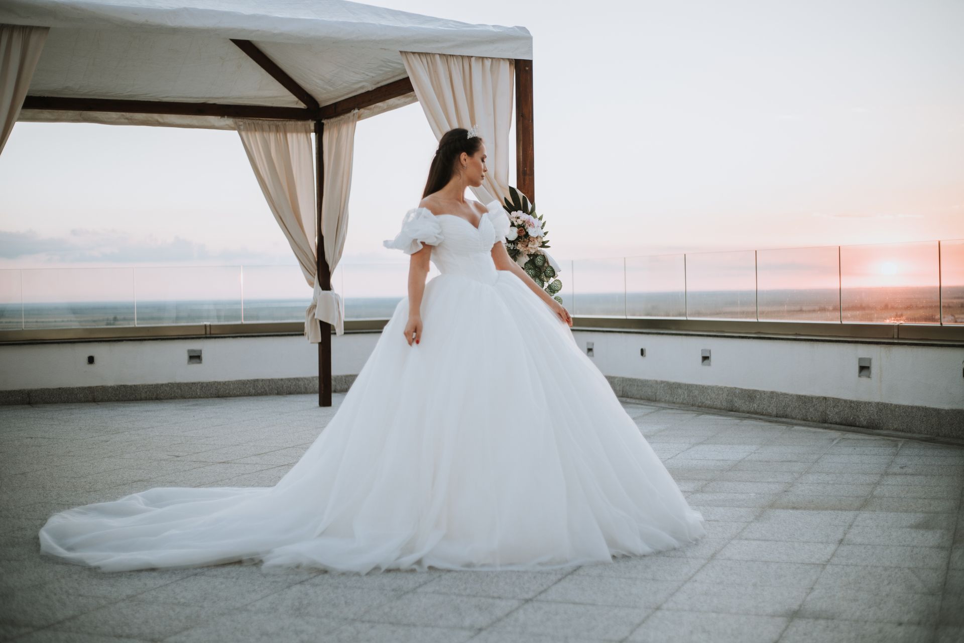 A bride in a wedding dress is standing on a balcony holding a bouquet of flowers.