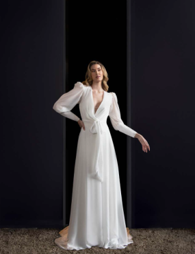 A woman in a white dress with long sleeves is standing in a doorway.