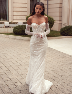 A woman in a white wedding dress and gloves is standing on a brick sidewalk.