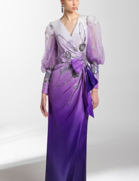 A woman in a long purple dress is standing in front of a white wall.