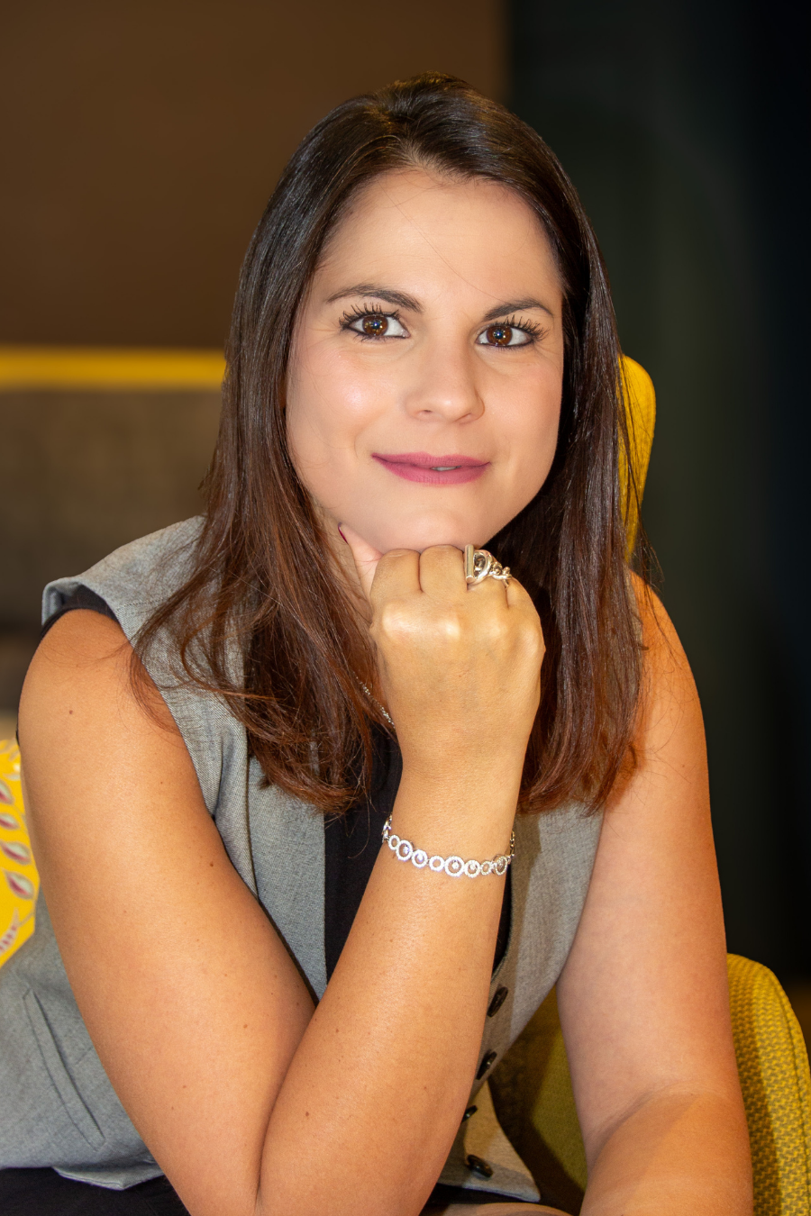 Une personne aux cheveux foncés, le menton appuyé sur sa main, porte un gilet gris sur un haut noir et est assise sur une chaise jaune.