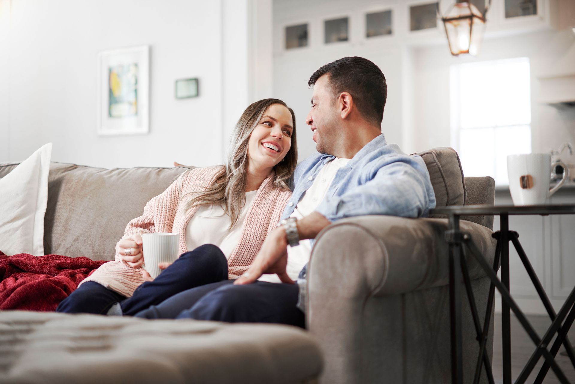 Husband and wife sitting on the living room couch drinking coffee and smiling at each other.