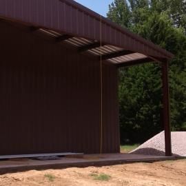 A dark brown metal building features an open porch area with a corrugated roof, a concrete slab, and a pile of gravel.