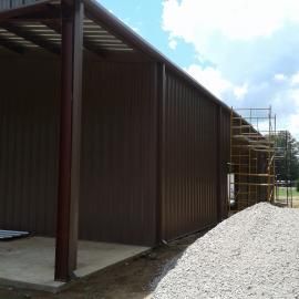 A brown metal warehouse under construction with scaffolding alongside a gravel pile against a blue sky with clouds.