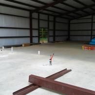 Interior of a metal building under construction with exposed steel beams, concrete floors, and plumbing pipe stubs.