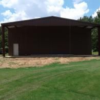 A brown metal barn or workshop with a single white door, situated on a patch of dirt in a grassy field under a cloudy sky.