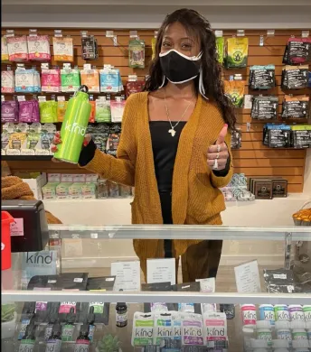Woman in mustard cardigan at a counter in a store, holding a green bottle and giving a thumbs up.