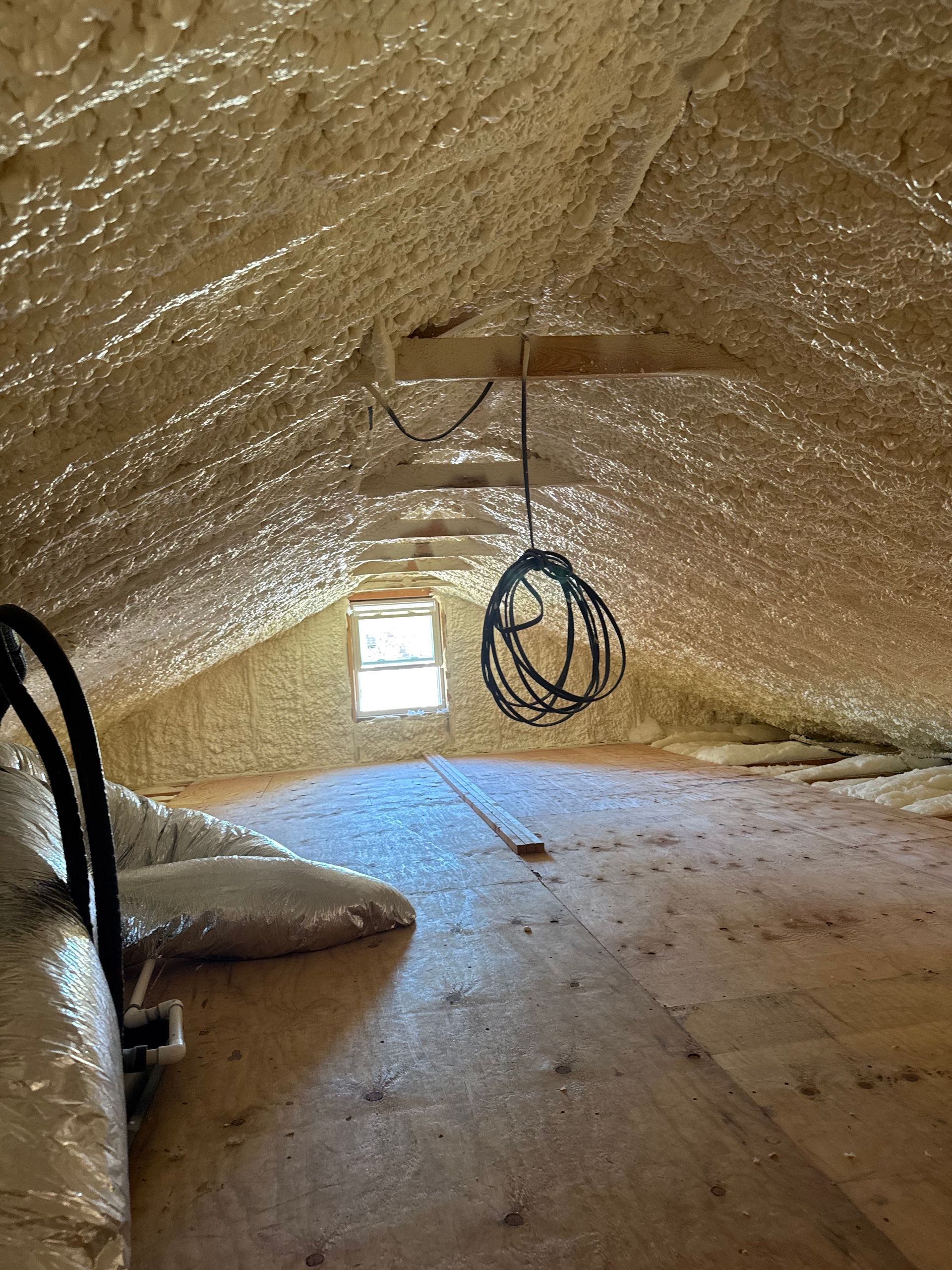 Attic interior with spray foam insulation on roof, wooden floor, small window, and hanging wires.