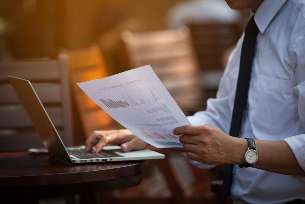 A Man Is Sitting at A Table Using a Laptop and Holding a Piece of Paper — Sunshine Coast Financial Advisers in Maroochydore, QLD