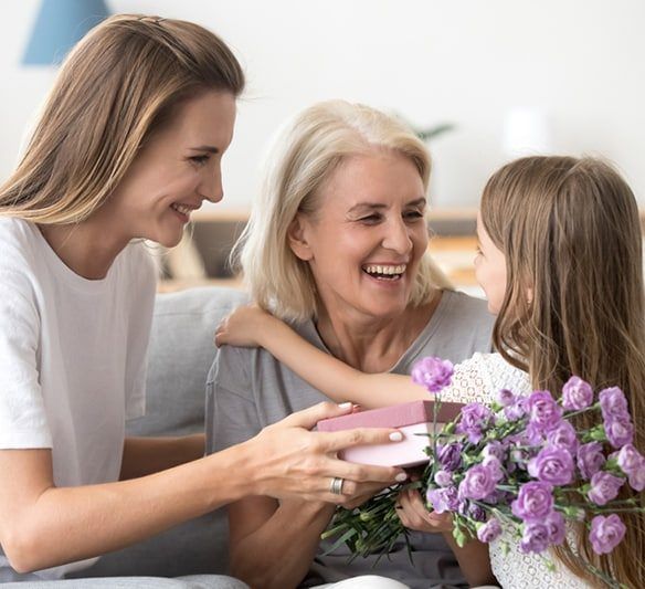 Two Women and A Little Girl Are Sitting on A Couch Holding Purple Flowers and A Gift Box — Sunshine Coast Financial Advisers in Maroochydore, QLD