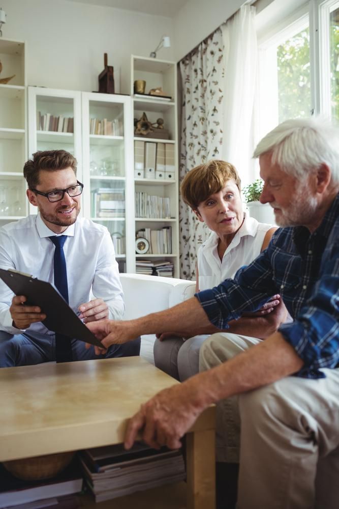 A Man Is Holding a Clipboard While Talking to An Elderly Couple in A Living Room — Sunshine Coast Financial Advisers in Maroochydore, QLD