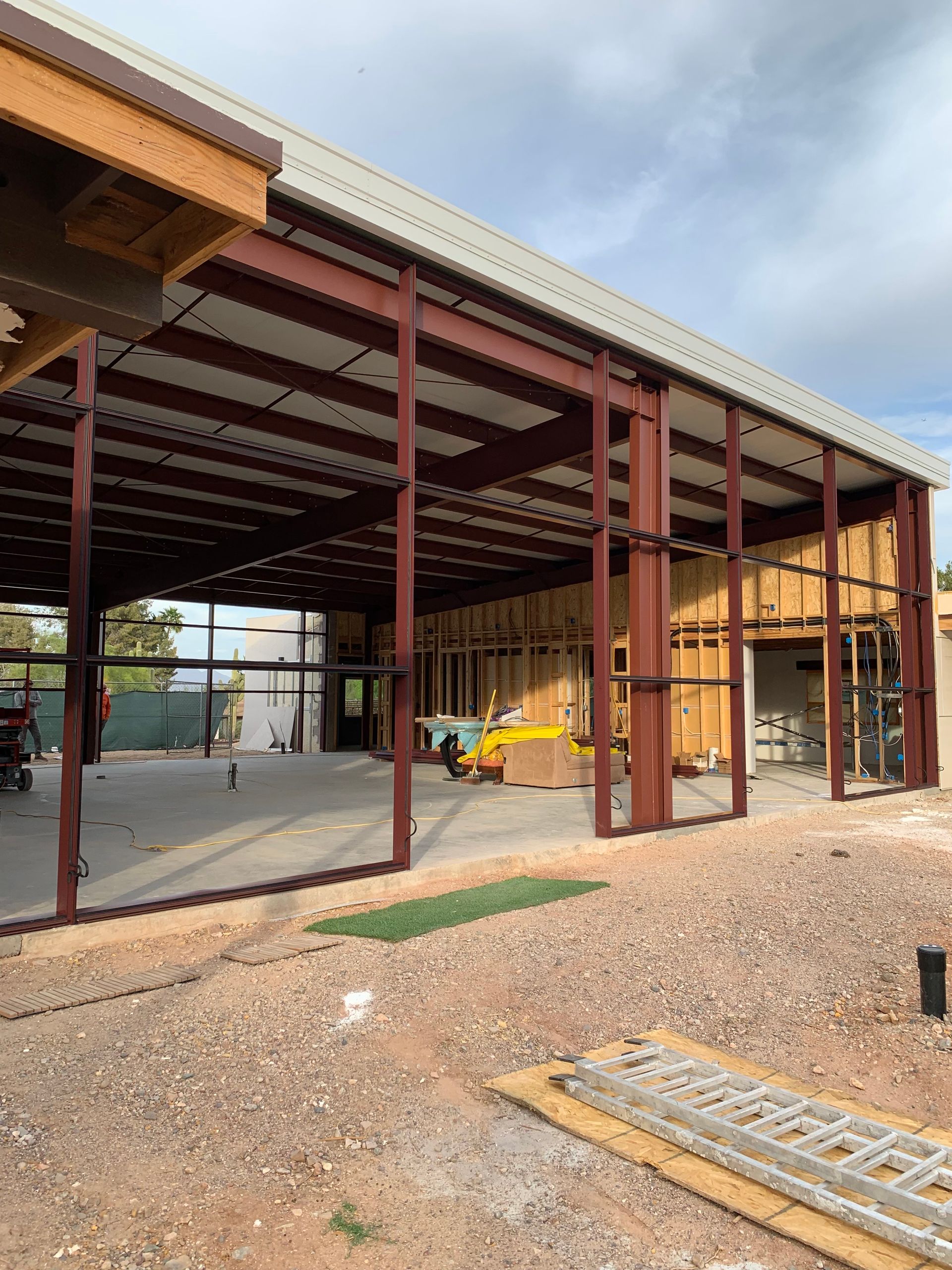 Steel-framed building under construction with exposed beams. Red steel, light roof, and dirt ground.