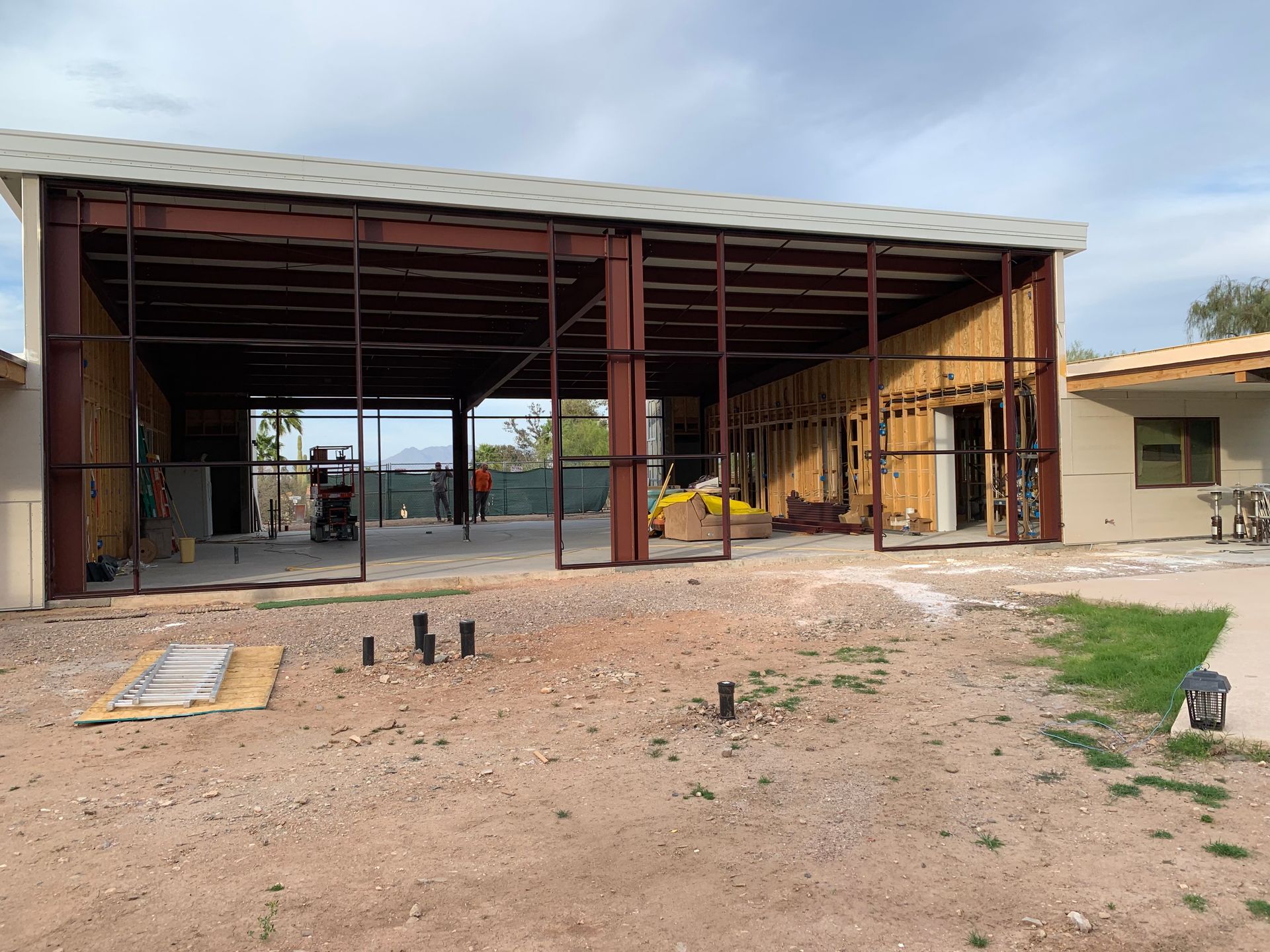 Steel-framed structure under construction, with open garage bays, adjacent to a residential building, and outdoor setting.