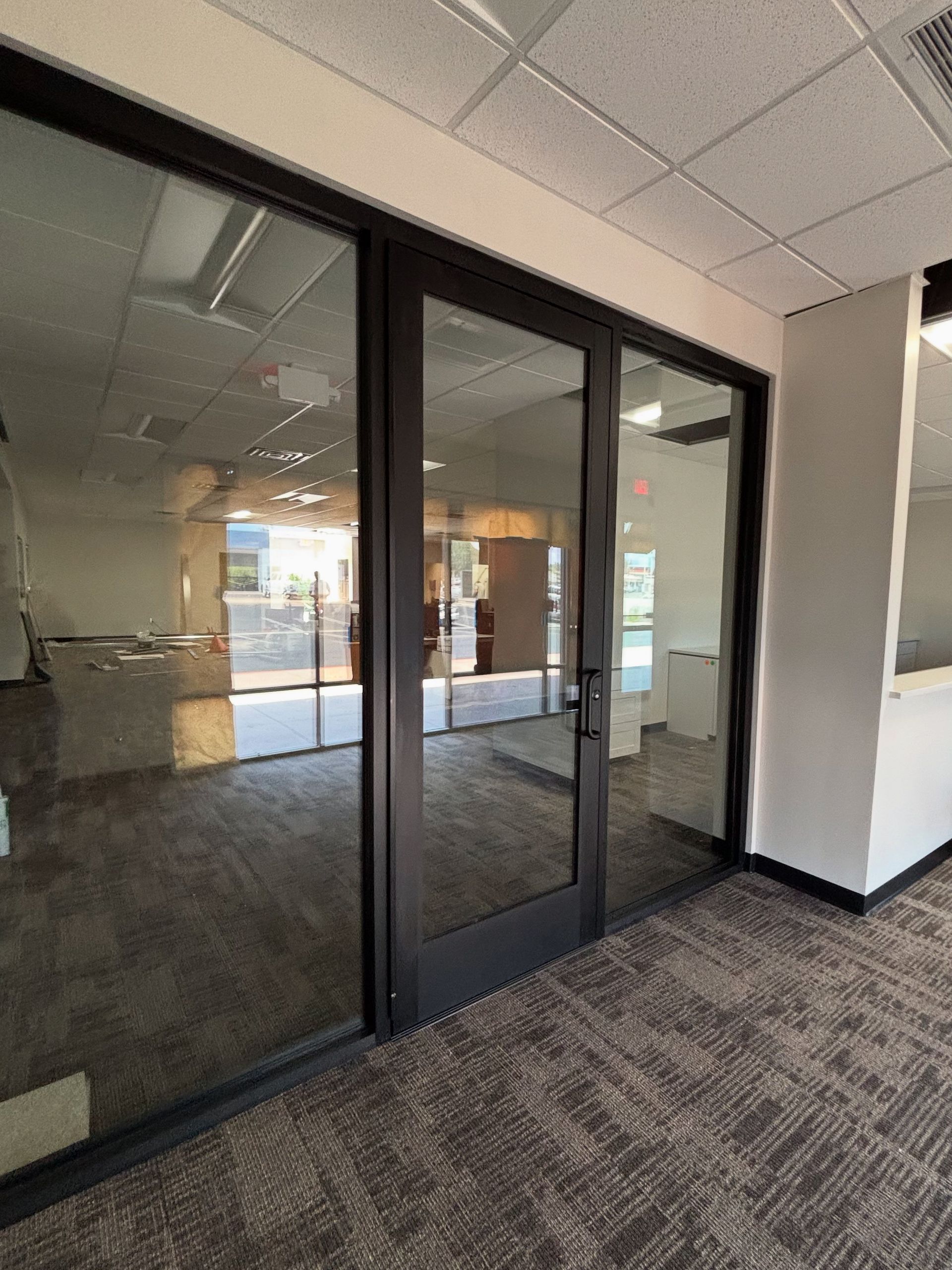 Glass double doors with dark frames, reflecting an empty interior and a hallway with patterned carpet.