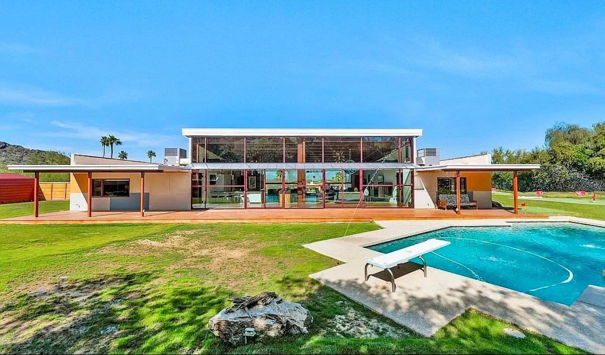 Mid-century modern house with large windows, pool, and green lawn under a blue sky.