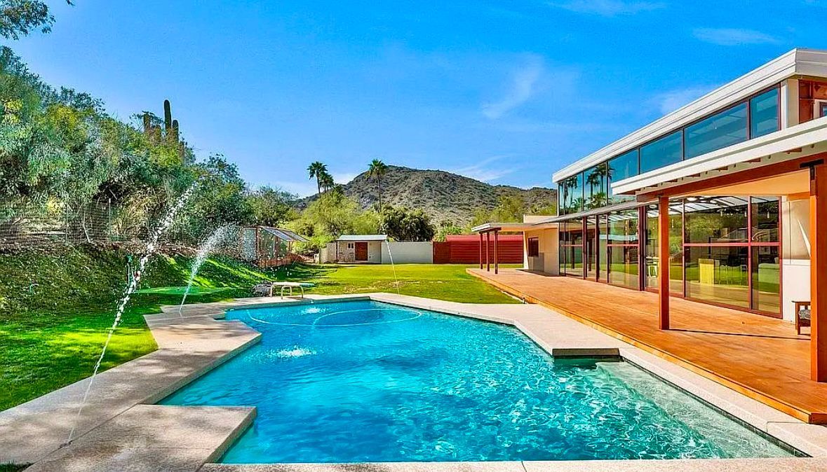 Swimming pool with house, lush green lawn, trees, and mountain backdrop under blue sky.