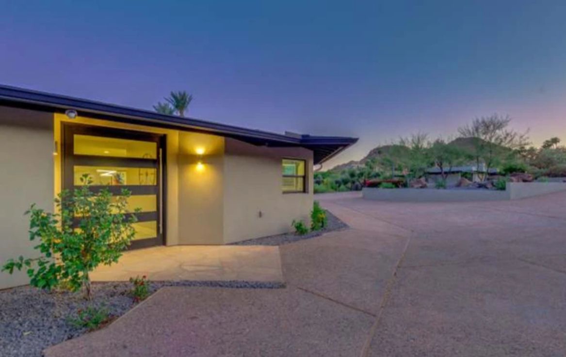Modern home exterior at dusk, tan stucco, dark frame entry door with glass panels. Driveway and desert landscape.