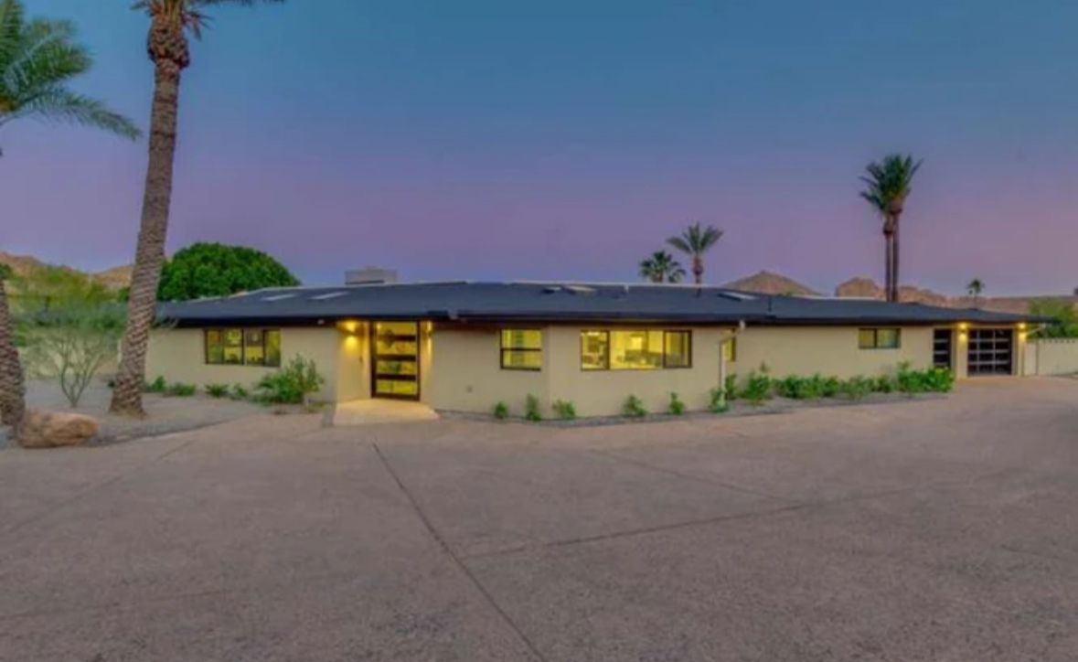 Beige single-story home with black roof, palm trees, and driveway under a dusky sky.