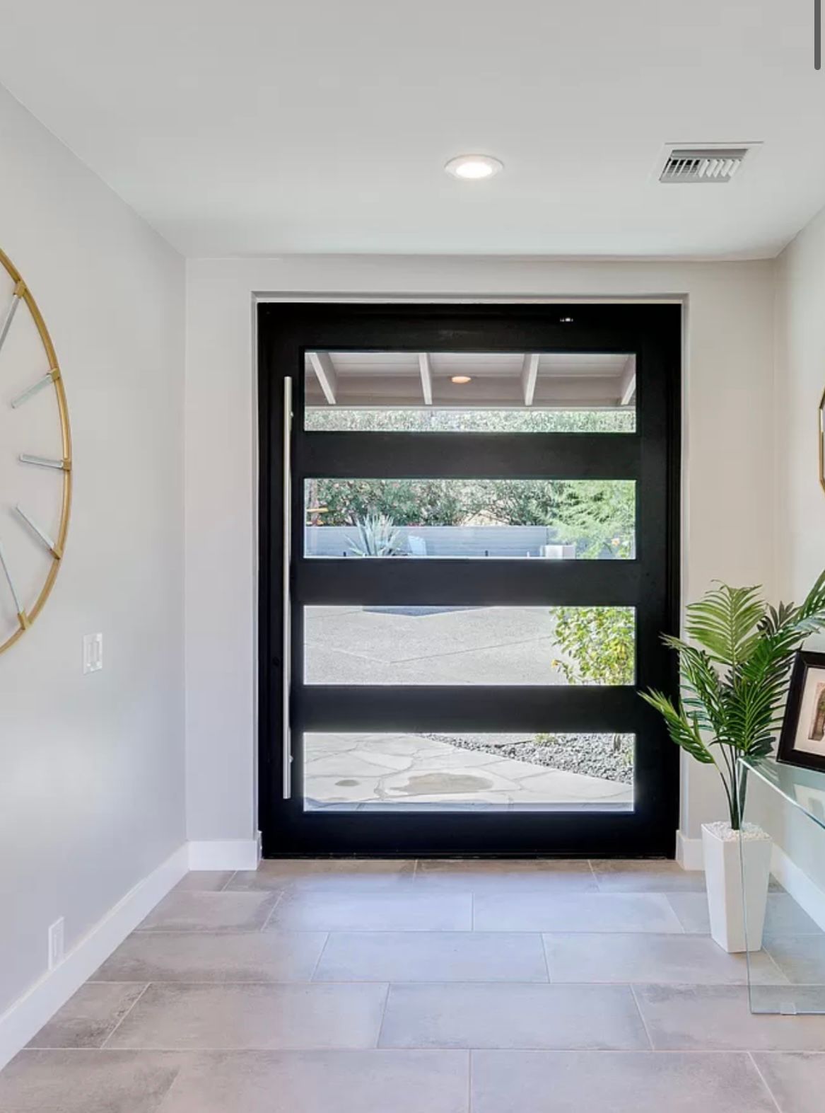 Modern black front door with horizontal glass panels. Interior view, white walls, large clock on left, plant on right.