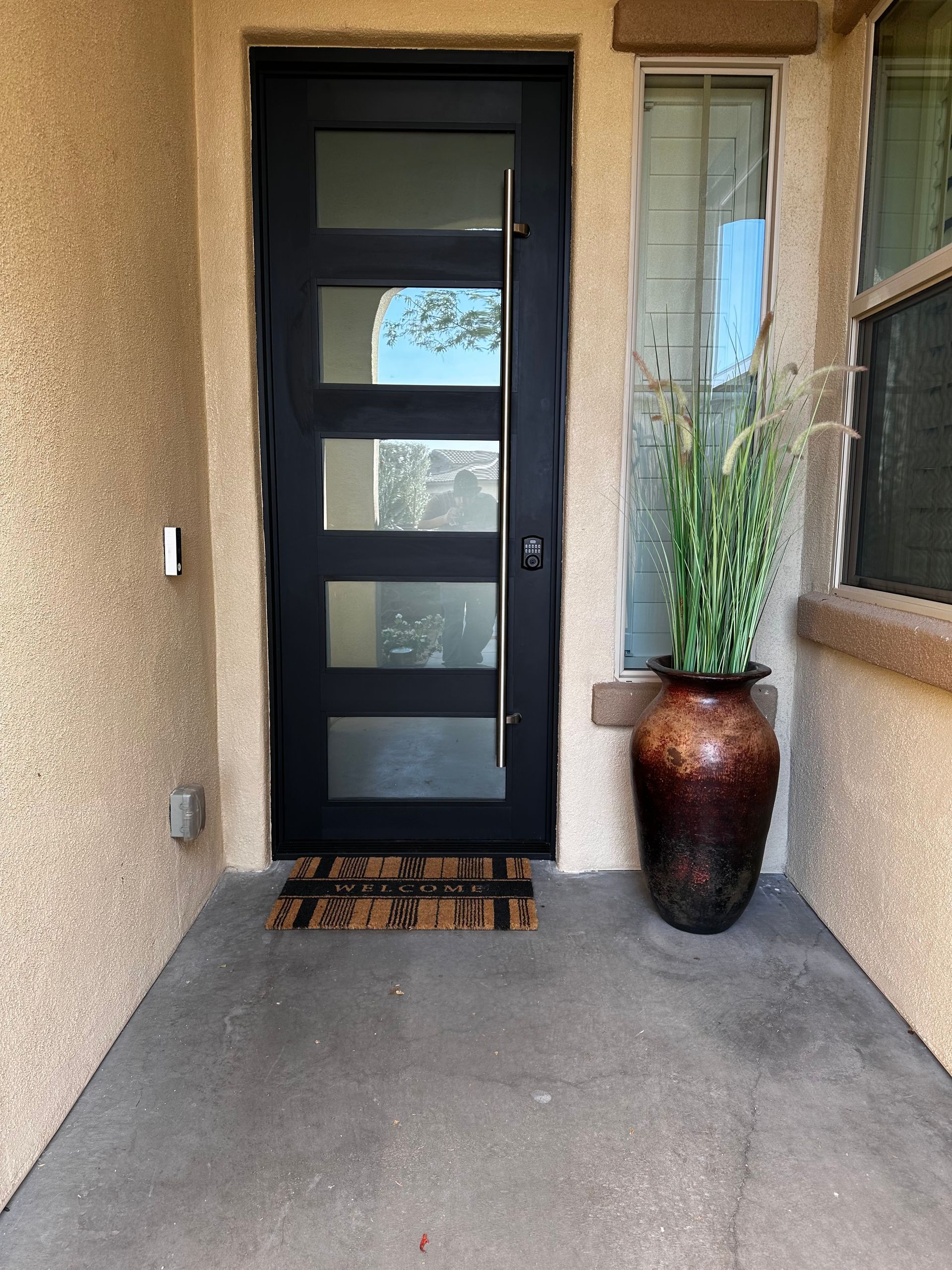 Black front door with four glass panels, a long handle, and a large brown planter on a porch.
