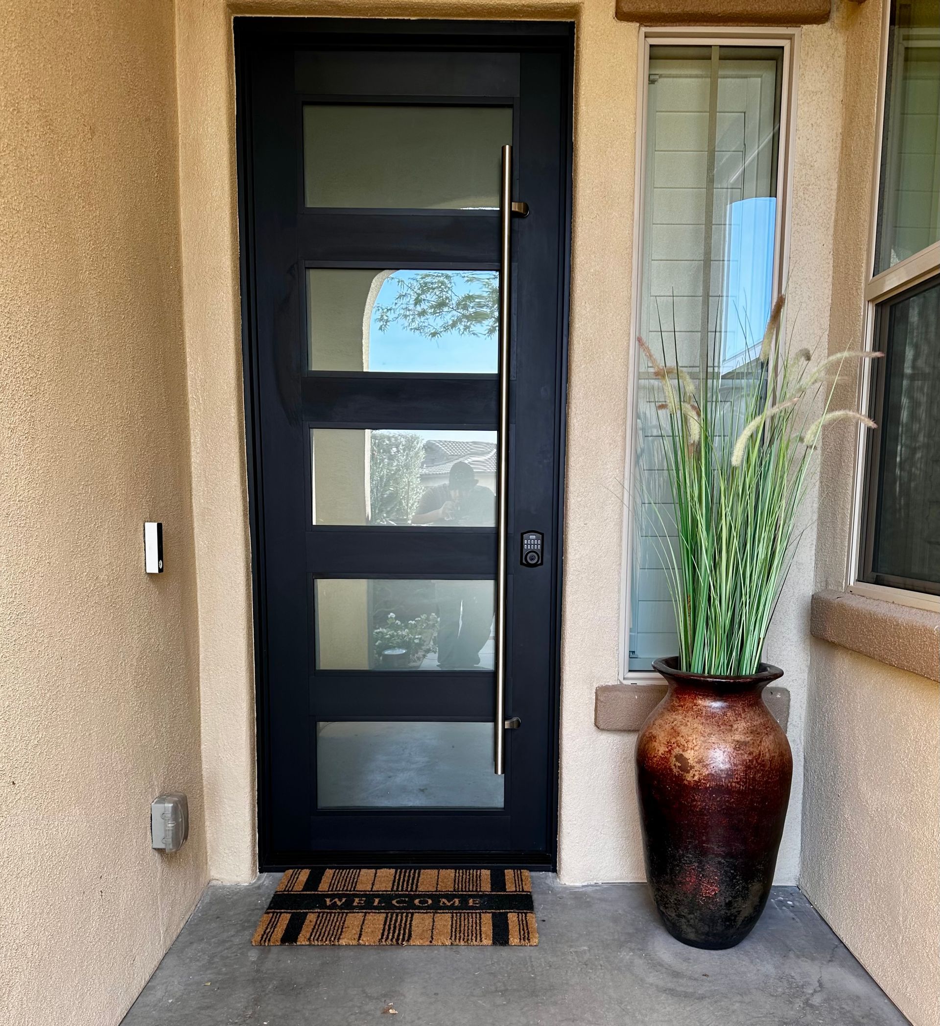 Black front door with four glass panels, long silver handle, welcome mat, and large brown pot with tall grass.