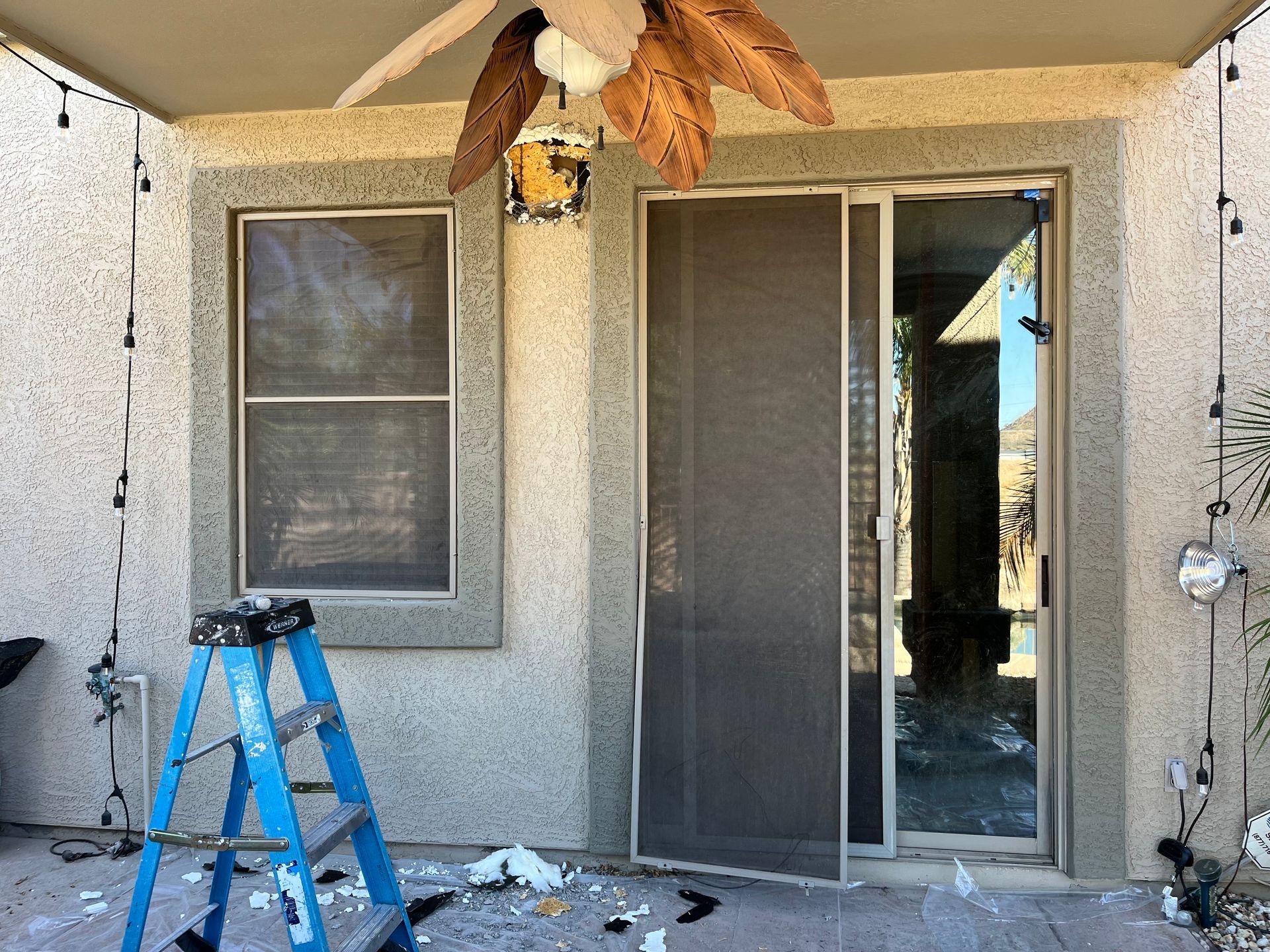 Exterior view of a stucco house with a sliding glass door and a window with a screen. A blue ladder is next to the window.