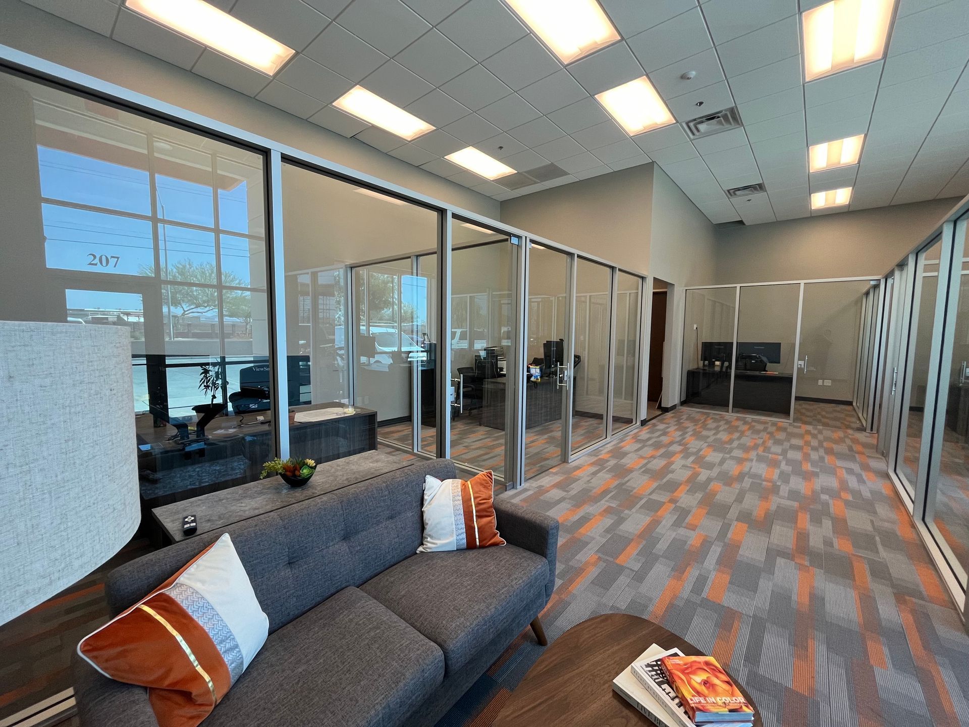 Waiting area with gray sofa, patterned carpet, glass-walled offices, and overhead lighting.