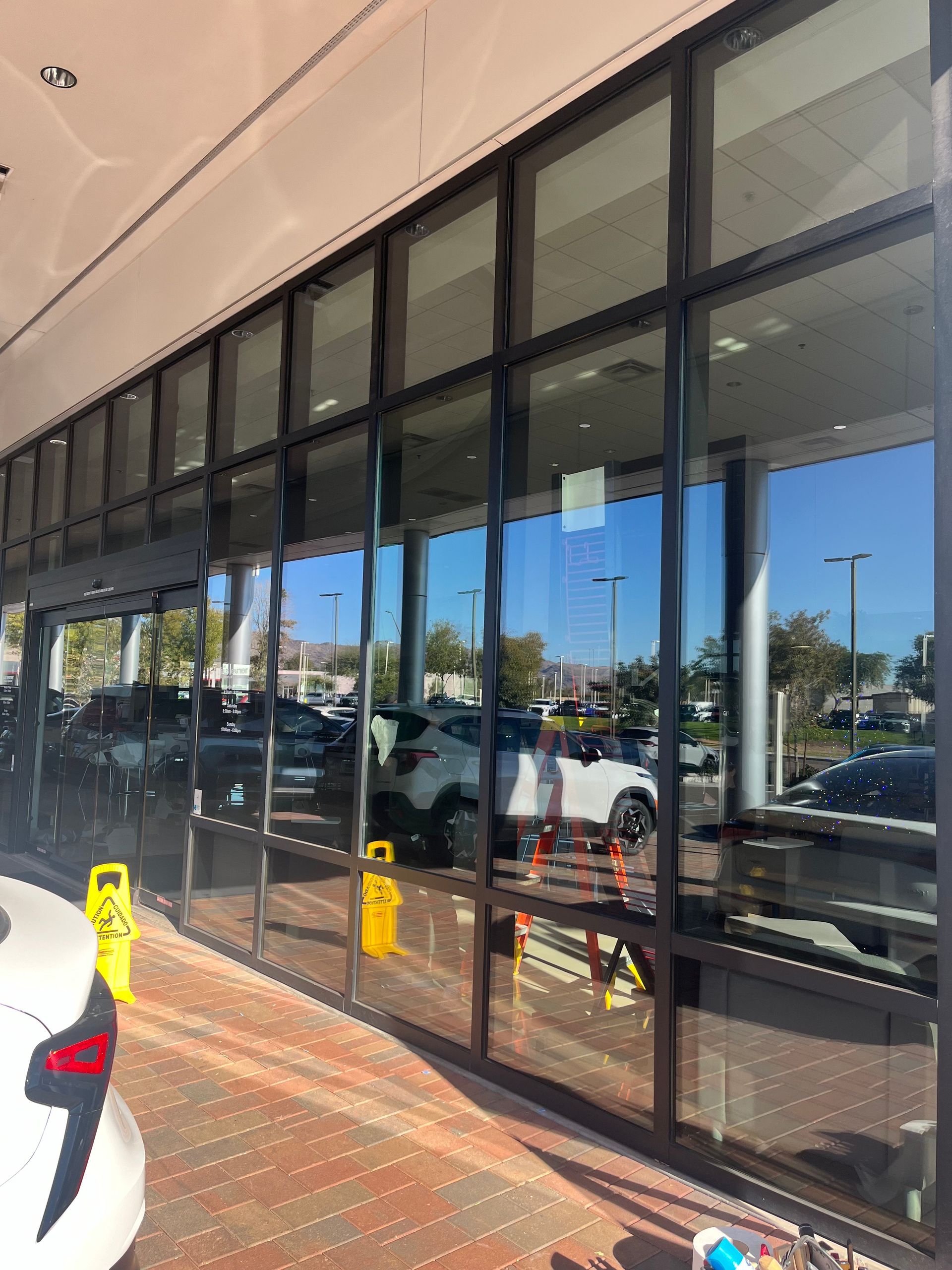 Glass storefront with black frames reflects cars and a clear sky; two yellow caution signs are visible.