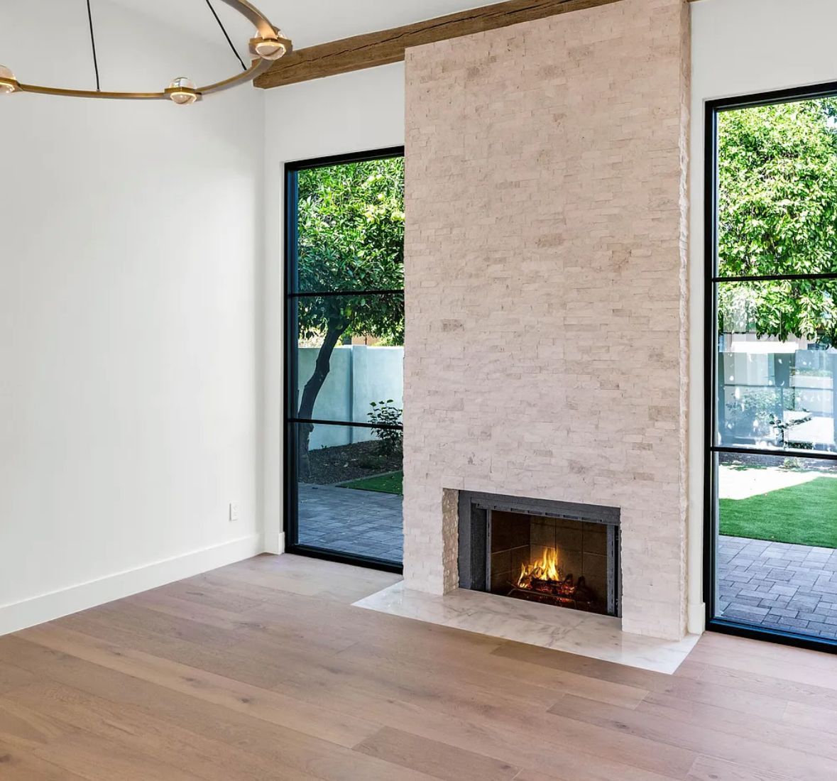 Modern living room with fireplace between two windows overlooking a backyard. Light wood floor and neutral walls.