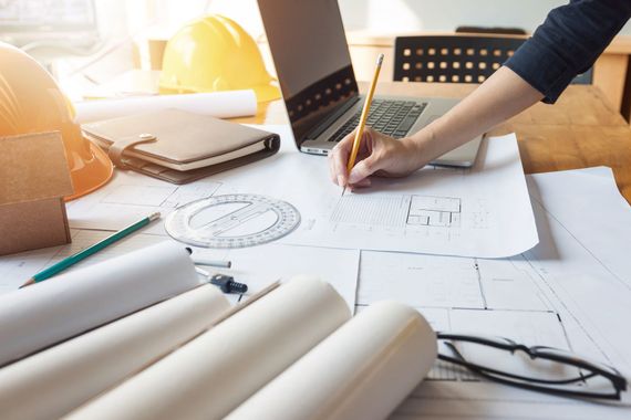 Person sketching on architectural blueprints at a sunlit desk with a laptop and hard hats Person sketching on architectural blueprints at a sunlit desk with a laptop and hard hats