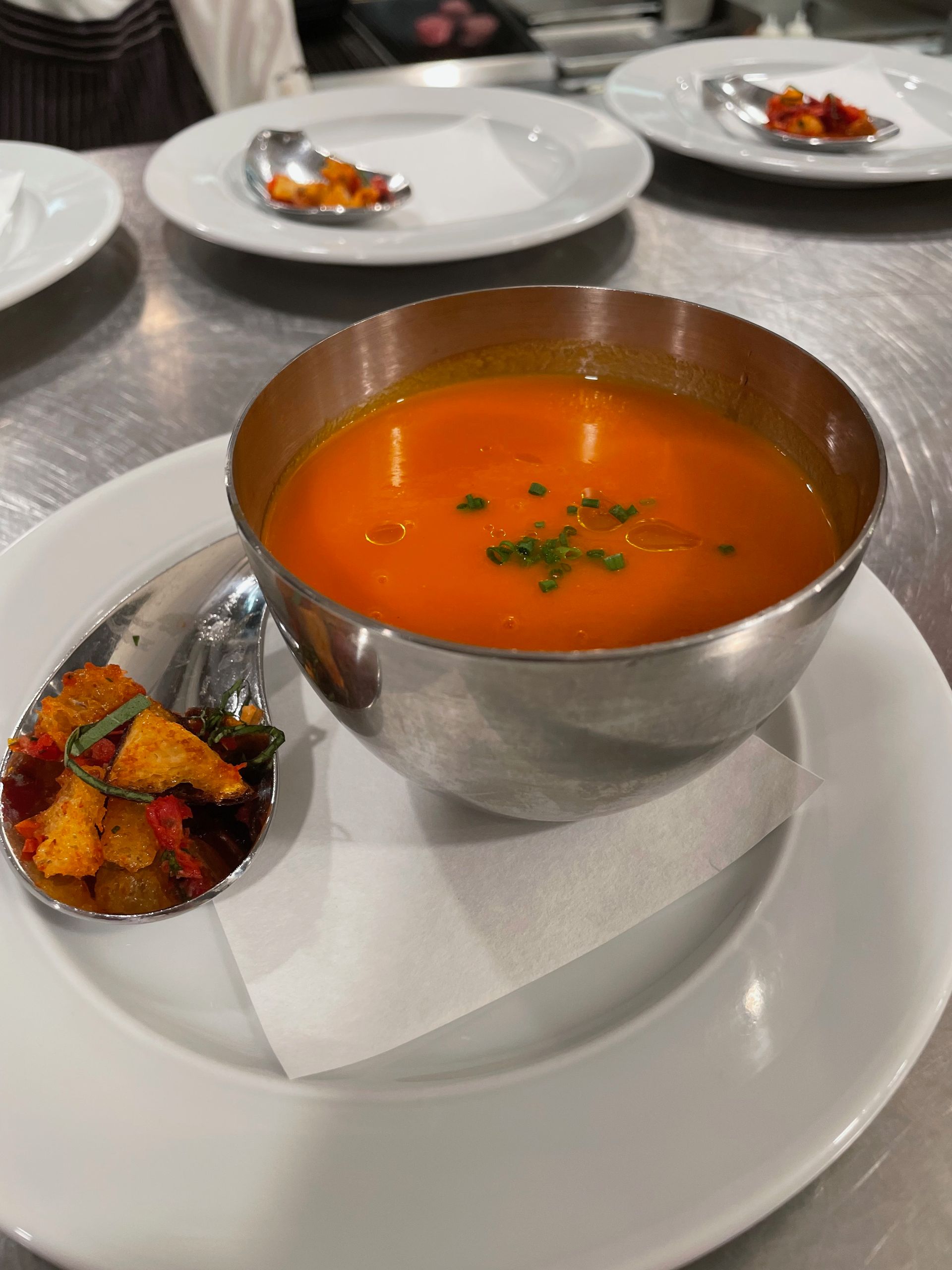 A bowl of orange soup with a spoon of colorful croutons on a white plate.