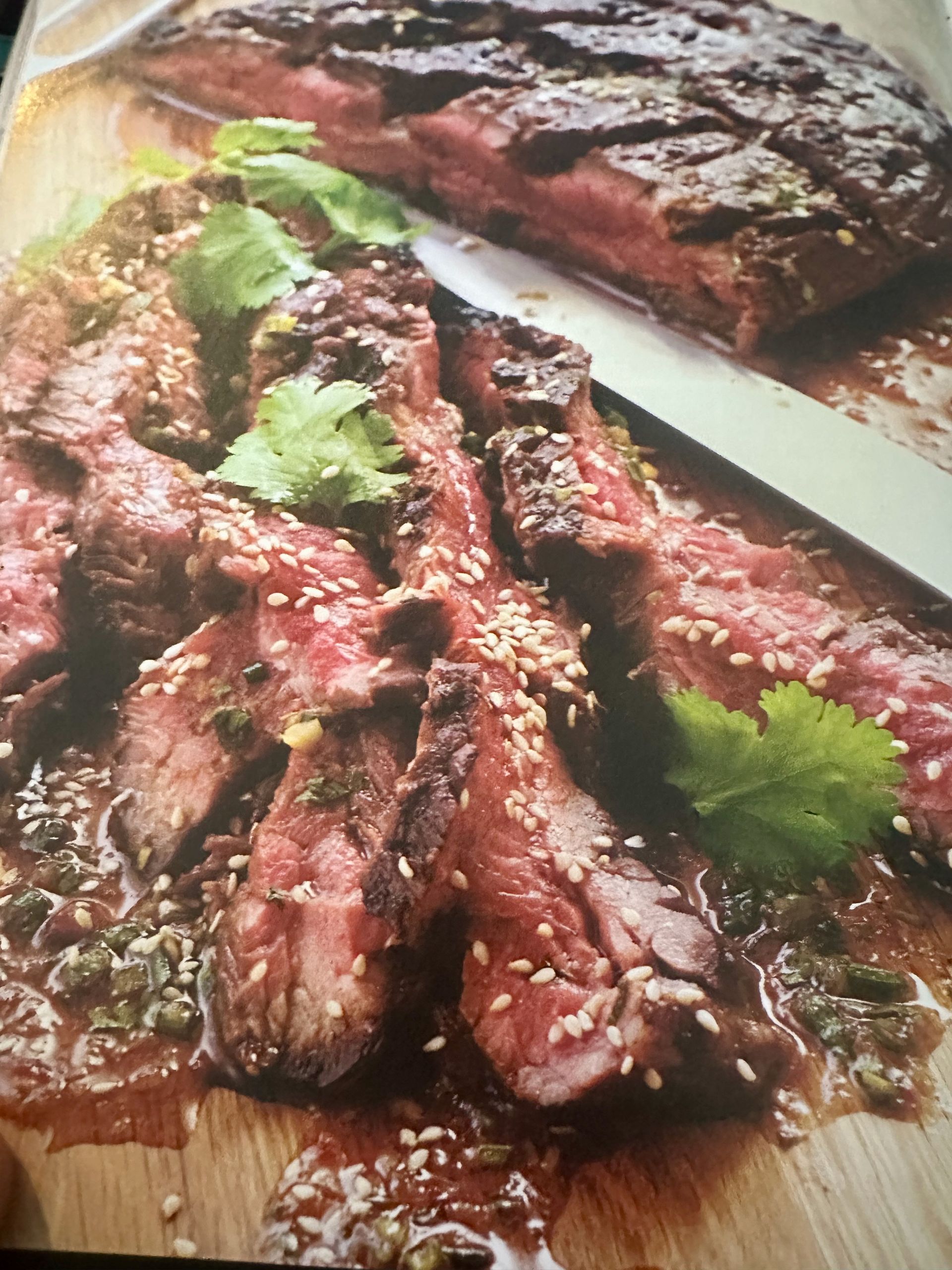 Sliced steak with sauce, sesame seeds, and cilantro on a wooden board.