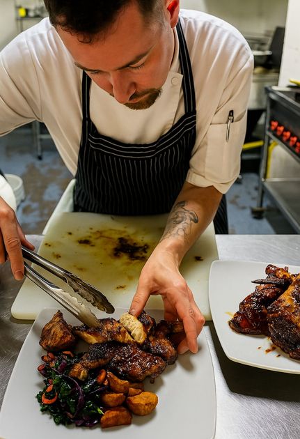 Chef plating food with tongs in a kitchen, dark-haired man wearing chef coat and apron, two plates visible.