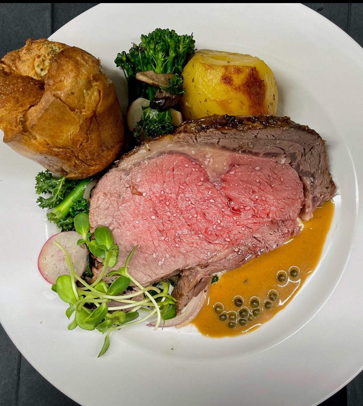 Roasted beef with Yorkshire pudding, vegetables, and sauce on a white plate.